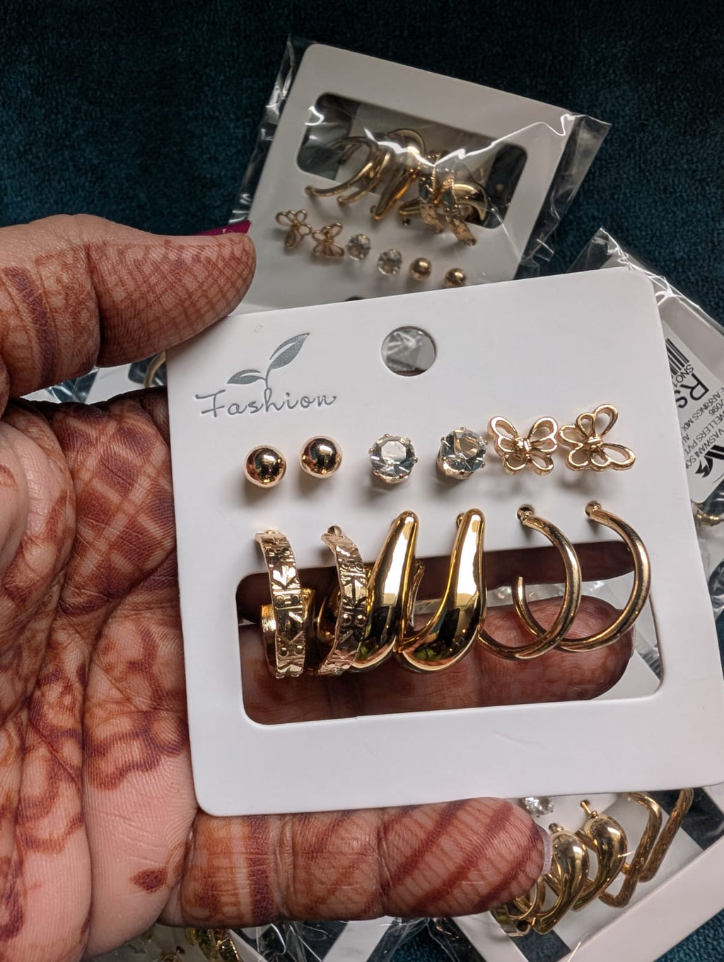 A hand with henna holding a white card displaying a set of gold and silver stud earrings and hoop earrings