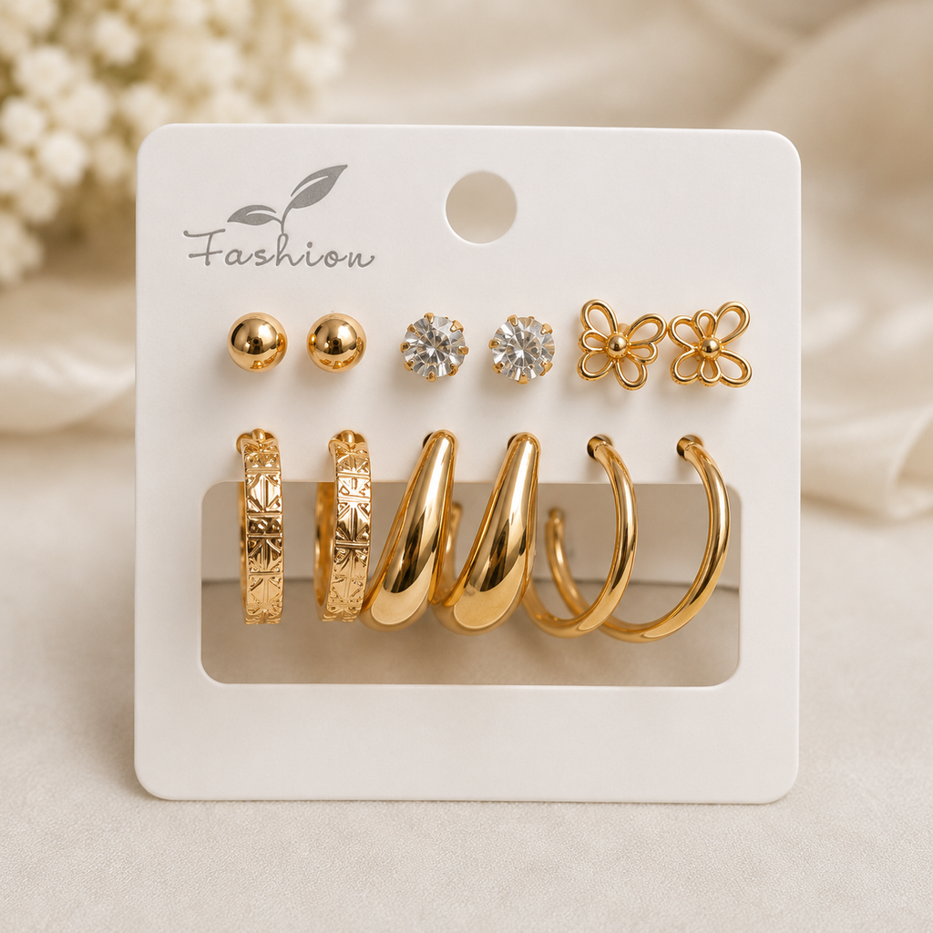 A set of gold-toned fashion earrings displayed on a white card, including two ball studs, two clear stone stud earrings, two flower-shaped stud earrings, and four huggie hoop earrings in different textures, on a soft beige background.