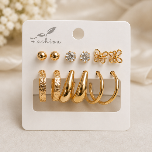 A set of gold-toned fashion earrings displayed on a white card, including two ball studs, two clear stone stud earrings, two flower-shaped stud earrings, and four huggie hoop earrings in different textures, on a soft beige background.